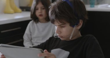 Ağız telleri olan bir çocuk mutfak masasında otururken dijital tabletine dalmış, yanında kahvaltı yapan başka bir çocuk gibi..