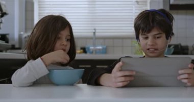 Ağız telleri olan bir çocuk mutfak masasında otururken dijital tabletine dalmış, yanında kahvaltı yapan başka bir çocuk gibi..