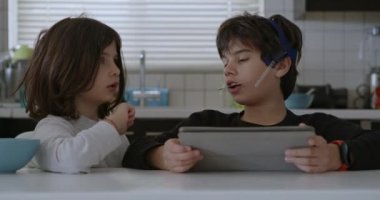 Ağız telleri olan bir çocuk mutfak masasında otururken dijital tabletine dalmış, yanında kahvaltı yapan başka bir çocuk gibi..