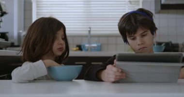 Ağız telleri olan bir çocuk mutfak masasında otururken dijital tabletine dalmış, yanında kahvaltı yapan başka bir çocuk gibi..