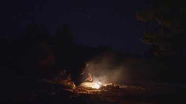 Karla çevrili bir kamp ateşiyle ellerini ısıtan bir adam görülüyor. Karanlık gökyüzünün altında..