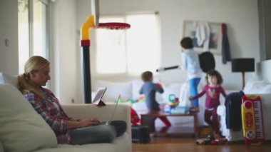 Çocukları oyuncaklarla dolu oturma odasında enerjik bir şekilde oynarken bir anne laptopuyla koltukta çalışır..