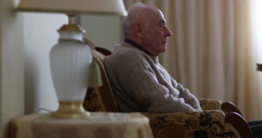 Yaşlı bir beyefendi, sakin bir yaşam alanına sıcak bir parıltı saçan bir lambayla yan manzaralı bir koltukta düşünceli bir şekilde oturur..