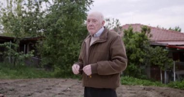 Sıcak bir ceket ve süveter giymiş düşünceli yaşlı bir adam solan bir akşam gökyüzünün zemininde kameraya bakıyor..