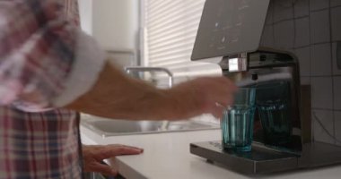 Bir adam modern bir filtreleme cihazından temiz bir bardağa su döküyor ve iyi ışıklandırılmış mutfağında sağlıklı bir yaşam tarzı seçtiğini vurguluyor..