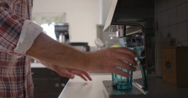 Bir adam mutfak tezgahında duruyor, modern bir ev suyu filtreleme sisteminin mavi bardağına kristal berrak su döküyor, doğal ışık sahneyi aydınlatıyor..
