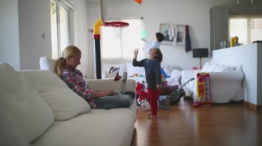 Odaklanmış bir anne, evinde çocuklarla uzaktan çalışmanın zorluklarını tasvir ederek, oyun oynayan çocukları etrafındaki oyunlarla uğraşırken, kanepede dizüstü bilgisayarıyla çalışır..