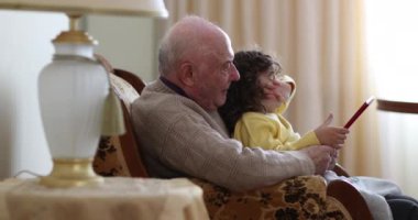 Bir büyükbaba ve küçük torunu, dijital tablete sarılmış bir şekilde oturma odasında birlikte oturuyorlar. Büyükbaba bej bir kazak giyer, sarı elbiseli çocuk ise ekrana odaklanır..
