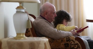 Bir büyükbaba ve küçük torunu, dijital tablete sarılmış bir şekilde oturma odasında birlikte oturuyorlar. Büyükbaba bej bir kazak giyer, sarı elbiseli çocuk ise ekrana odaklanır..