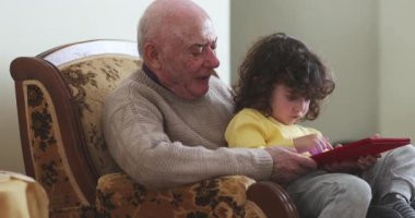 Bir büyükbaba ve küçük torunu, dijital tablete sarılmış bir şekilde oturma odasında birlikte oturuyorlar. Büyükbaba bej bir kazak giyer, sarı elbiseli çocuk ise ekrana odaklanır..
