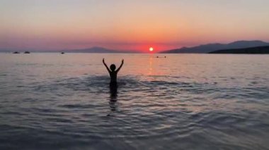 Genç bir çocuk gün batımında bir plajın yumuşak dalgalarında oynar. Güneş uzak dağların üzerinden batarken kollarını neşeyle kaldırıyor, sakin denizin üzerine sıcak bir parıltı saçıyor..