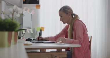 Sarışın bir kadın, monitöründeki bitkiler ve yapışkan notlarla çevrili ev ofisindeki uzaktan çalışmaya odaklanmış..