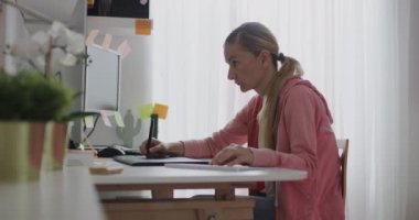Sarışın bir kadın, monitöründeki bitkiler ve yapışkan notlarla çevrili ev ofisindeki uzaktan çalışmaya odaklanmış..