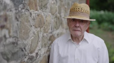 Beyaz gömlekli ve hasır şapkalı yaşlı bir adam kırsal taştan bir duvara bakıyor ve doğrudan kameraya bakıyor..