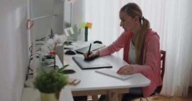 Bir kadın, gün boyunca iyi aydınlatılmış bir odada ofis malzemeleriyle çevrili bir masada dijital tablet ve bilgisayar kullanarak uzaktan çalışıyor..