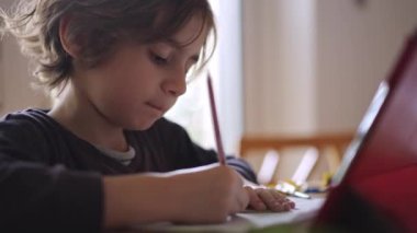 Odaklanmış genç bir çocuk, iyi ışıklandırılmış bir odada bir masada otururken dijital tabletine dikkatle çizer..