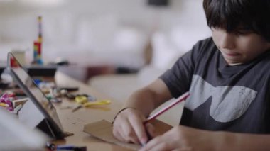 Genç bir çocuk, iyi aydınlatılmış bir odadaki bir masadaki dijital tablet üzerindeki talimatlara atıfta bulunarak bir kağıt işi projesine konsantre olur..