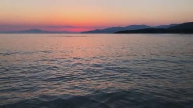 Çarpıcı bir günbatımı, sakin bir denizin üzerine canlı renkler saçar. Arka planda dağlar, alacakaranlıkta sakin bir plaj sahnesi oluşturur..