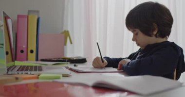 Donanma kapüşonlusu giyen genç bir çocuk, sabit malzemelerle çevrili bir dizüstü bilgisayar kullanarak bir masada özenle ödevlerini yapar..
