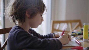 Genç bir çocuk, iyi aydınlatılmış bir odadaki bir masadaki dijital tablet üzerindeki talimatlara atıfta bulunarak bir kağıt işi projesine konsantre olur..