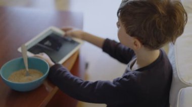 Bir çocuk, sıcak bir oturma odasında oturmuş bir kaseden kahvaltı ederken dijital tabletle meşgul oluyor..