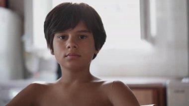 Kısa siyah saçlı bir çocuk doğrudan kameraya bakıyor. Bir odadaki pencerelerden süzülen güneş ışığıyla aydınlatılıyor..