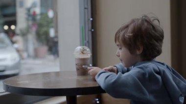 Akıllı telefonuna kendini kaptırmış genç bir çocuk bir kahve dükkanının penceresinde masada bir içecekle oturuyor..