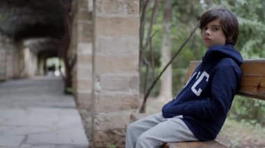 Ciddi bir ifadeye sahip genç bir çocuk parktaki ahşap bir bankta oturuyor, elleri kapüşonlu cebinde, ağaçlarla çevrili..
