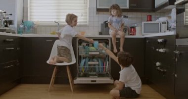 Gündelik kıyafetli üç çocuk, bulaşıkları bulaşık makinesine koyuyor, modern bir mutfakta takım çalışmasını ve iş birliğini teşvik ediyor..