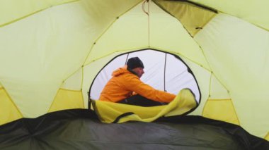 Bir adam sarı bir çadırda oturuyor, kışlık dağların ortasında, sıcacık giyinmiş, giysilerini düzenliyor..