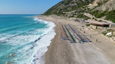 Yunanistan 'ın Lefkada kentinde kıyı boyunca sıralanmış güneş yatakları ve şemsiyeleri gösteren bir plaj manzarası. Turkuaz su kumlu kumsala çarpıyor ve güzel bir manzara yaratıyor..