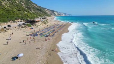 Yunanistan 'ın Lefkada kentinde kıyı boyunca sıralanmış güneş yatakları ve şemsiyeleri gösteren bir plaj manzarası. Turkuaz su kumlu kumsala çarpıyor ve güzel bir manzara yaratıyor..