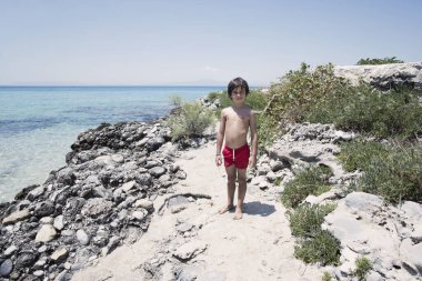 Kızıl mayo giymiş genç bir çocuk, güneşli bir günde, kayalık bir kıyı şeridinde dikilip, Akdeniz 'in sakin mavi sularına bakıyor..