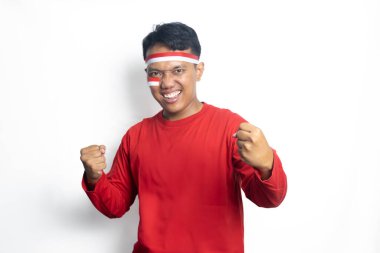 Endonezya 'nın kırmızı tişörtünü ve kırmızı bayrağını giymiş genç Asyalı bir adam beyaz arka planda mutlu bir gülümsemeyle zaferi zaferle kutluyor.