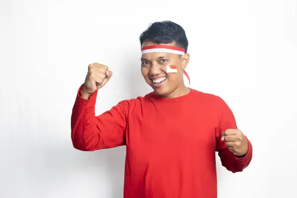 Endonezya 'nın kırmızı tişörtünü ve kırmızı bayrağını giymiş genç Asyalı bir adam beyaz arka planda mutlu bir gülümsemeyle zaferi zaferle kutluyor.