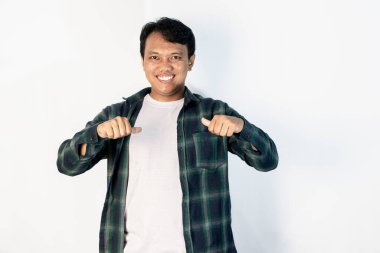 Yeşil tişörtlü, neşeli Güneydoğu Asyalı adam iki başparmağı da kendisine dönük şekilde gülümsüyor. Beyaz bir arkaplanda izole edilmiş.