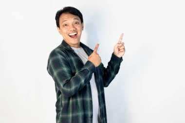 Yeşil gömlekli neşeli genç Asyalı adam, iki parmağıyla yukarıya doğru işaret ederken geniş bir gülümseme sergiliyor, beyaz arka planda güvenle duruyor..