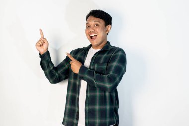 Yeşil gömlekli neşeli genç Asyalı adam, iki parmağıyla yukarıya doğru işaret ederken geniş bir gülümseme sergiliyor, beyaz arka planda güvenle duruyor..