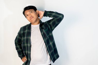 Asyalı yeşil gömlekli bir adam kafasının arkasını kaşıyor. Kafası karışık ve temel bir ifadeyle. Beyaz bir stüdyonun arka planında duruyor.