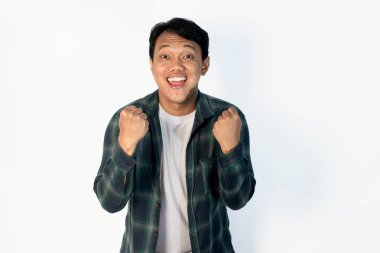 Yeşil tişörtlü genç Asyalı adam, beyaz arka planda çok mutlu ve heyecanlı bir şekilde kolları havada, gülümseyerek ve başarı çığlıkları atarak kazanan bir jest yapıyor. Kutlama