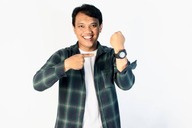 Beyaz arka planda sıradan bir gömlek giyen genç Asyalı adam saati işaret ediyor, mutlu, zamanı geldi, kameraya gülümseyerek bakıyor.