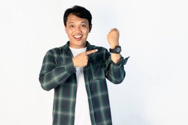 Beyaz arka planda sıradan bir gömlek giyen genç Asyalı adam saati işaret ediyor, mutlu, zamanı geldi, kameraya gülümseyerek bakıyor.