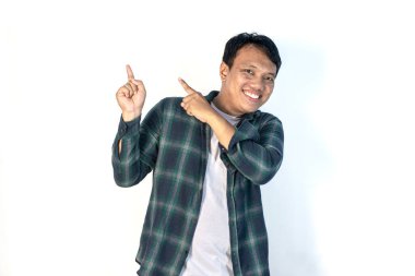 Yeşil gömlekli neşeli genç Asyalı adam, iki parmağıyla yukarıya doğru işaret ederken geniş bir gülümseme sergiliyor, beyaz arka planda güvenle duruyor..
