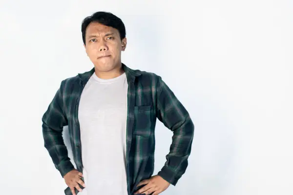 Genç Asyalı (Endonezyalı) adam beyaz arka planda, elleri kalçasında gezerken yüzü şüpheyle izole edilmiş.