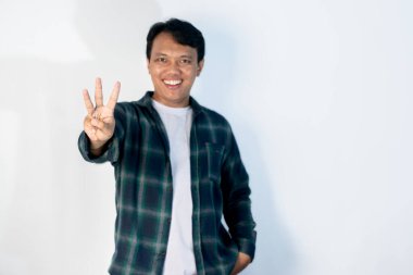 Yeşil tişörtlü genç Endonezyalı adam izole edilmiş beyaz arka planda duruyor ve üç numaralı parmaklarıyla işaret ederken kendinden emin ve mutlu bir şekilde gülümsüyor..