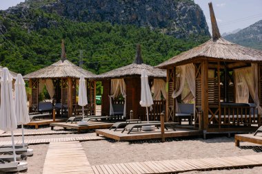 Güneşli şezlongları, çardakları, masaları ve beyaz şemsiyeleri olan güzel, boş bir kumsal dağların arkasında, yaz mevsiminde.