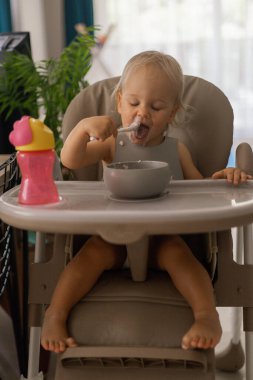 Küçük, güzel, sarışın bir kız, çocukların yemek masasında otururken kaşıkla yer.