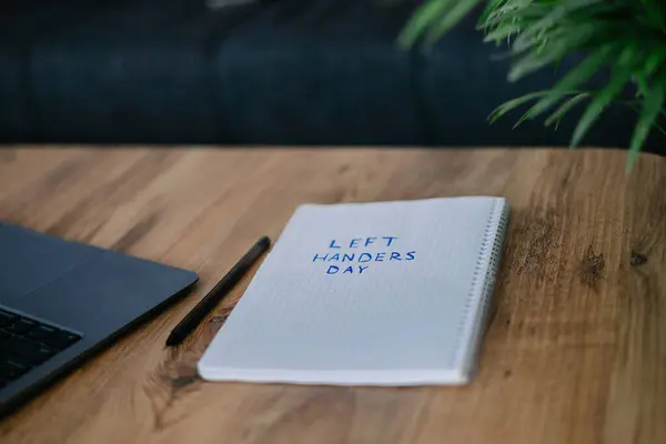 stock image a notebook with a text Left handers day on a wooden table. blured flower, black pen and laptop 