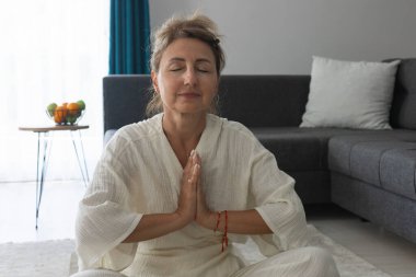 Beyaz elbiseli, orta yaşlı güzel bir kadın, evdeki bir odada gri bir halıda otururken egzersiz ve meditasyon yapıyor. Spor ve sağlık.