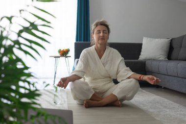 Beyaz elbiseli, orta yaşlı güzel bir kadın, evdeki bir odada gri bir halıda otururken egzersiz ve meditasyon yapıyor. Spor ve sağlık.